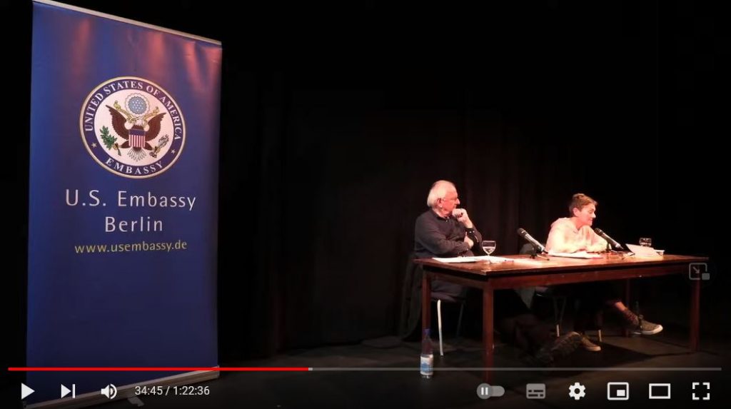 Screenshot from reading on stage: Two people sitting at a desk: The man is listening to a woman